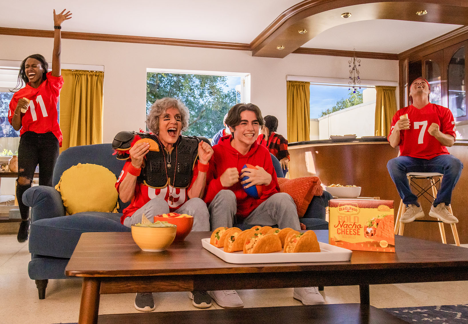 Family in the living room wearing football jerseys, watching a game, with tacos, chips, and a box of Old El Paso Bold Nacho Cheese taco Shells, front of package, on the table